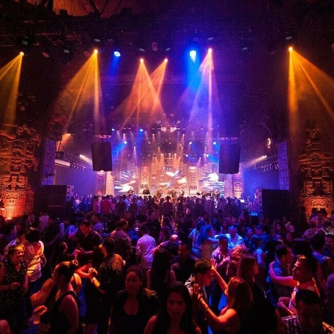 Crowd of people gathered on a dance floor facing a brightly lit stage with blue and amber concert lighting.