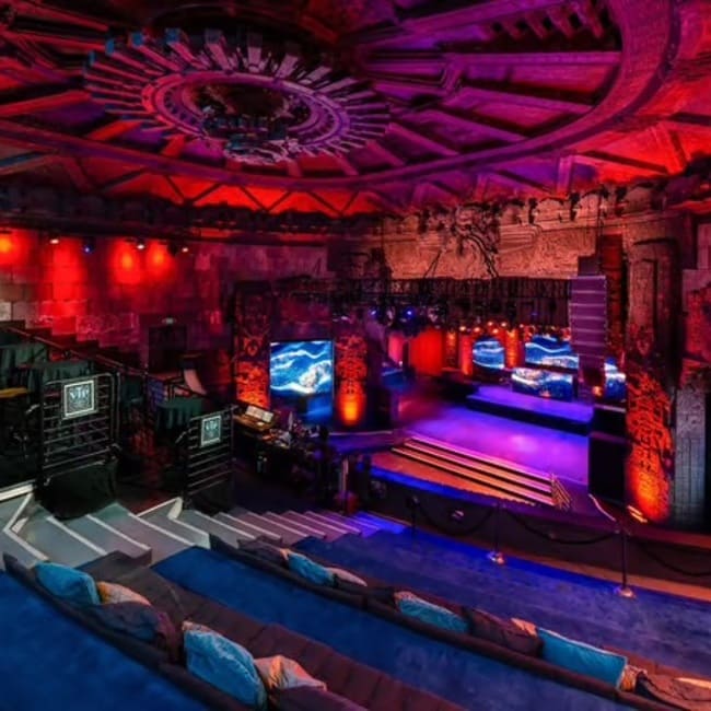 Elevated view of an indoor venue with tiered seating, a raised stage, steps, and dramatic red and purple lighting along carved walls and ceiling.